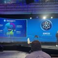 Speaker on stage at a conference with a large screen showing a blue 'Ideas Intelligence' presentation and a puzzle-piece graphic labeled trust.
