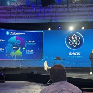 Speaker on stage at a conference with a large screen showing a blue 'Ideas Intelligence' presentation and a puzzle-piece graphic labeled trust.