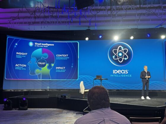 Speaker on stage at a conference with a large screen showing a blue 'Ideas Intelligence' presentation and a puzzle-piece graphic labeled trust.