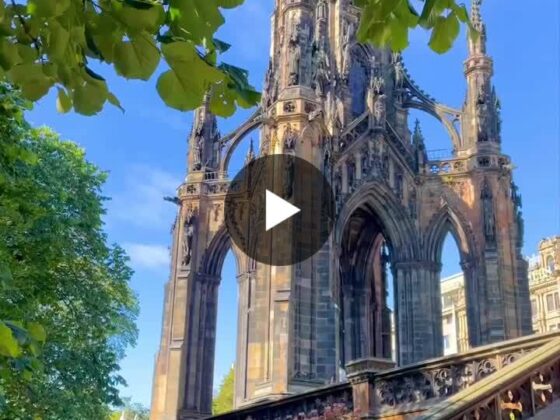 Gothic ruin with ornate arches framed by green leaves and a blue sky, stairs with decorative railing in the foreground.