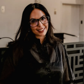 Smiling woman with dark hair and glasses standing in a modern office hallway. She wears a dark blouse and looks at the camera.