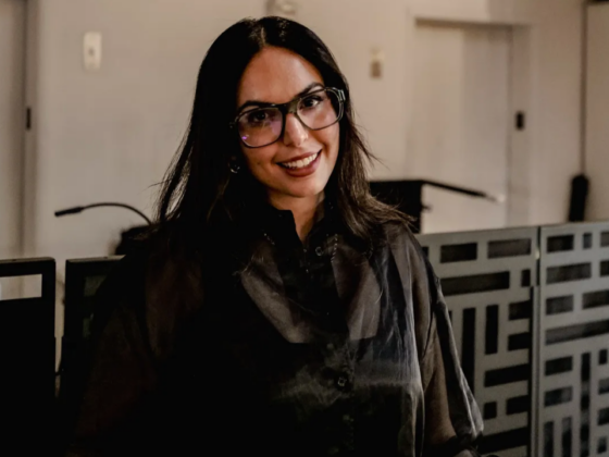 Smiling woman with dark hair and glasses standing in a modern office hallway. She wears a dark blouse and looks at the camera.