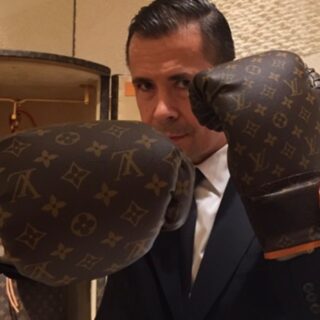 Man in a dark suit wearing oversized Louis Vuitton boxing gloves, posing with fists up in front of the camera in a luxury store." (informational)