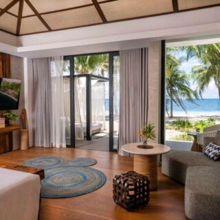 Luxurious hotel suite with a beach view through glass doors, palm trees outside, and a living area with sofa and TV.