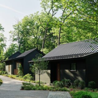 Row of dark wooden cabins along a winding gravel path in a lush green forest setting.