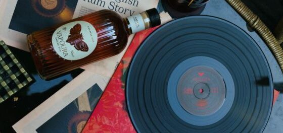 A vinyl record sits on a turntable with photos, papers, and a whiskey bottle scattered around on a cluttered table.