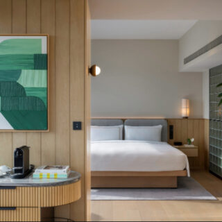 Hotel-style bedroom seen through a doorway: queen bed with grey headboard, wood-paneled walls, and a curved marble coffee station on the left with a coffee machine and books beneath.