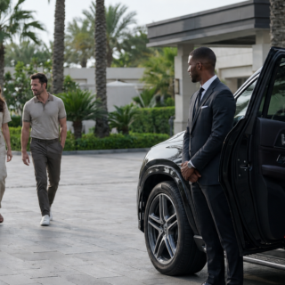 Chauffeur in a dark suit stands by an open black SUV as a family of three approaches at a luxury hotel entrance.
