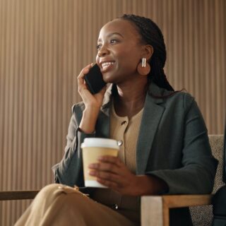 Professional woman smiles while talking on her phone, holding a coffee cup in a lobby with a suitcase beside her.