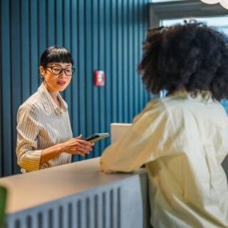 Receptionist with glasses assists guests at a modern front desk in a blue-walled lobby.