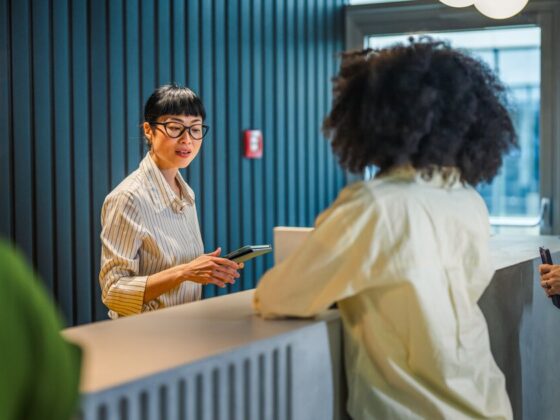 Receptionist with glasses assists guests at a modern front desk in a blue-walled lobby.