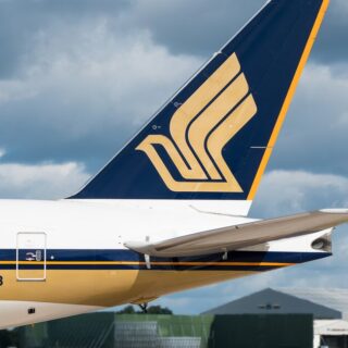 Close-up of a commercial airplane tail with a blue and gold logo, parked at an airport runway/hangar backdrop.