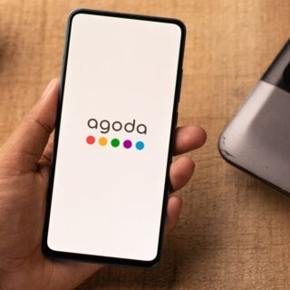 Phone in hand showing Agoda app logo on white screen, on a wooden desk near a laptop and glasses.