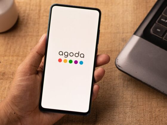 Phone in hand showing Agoda app logo on white screen, on a wooden desk near a laptop and glasses.