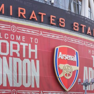 Exterior of Emirates Stadium with a large red banner reading 'Welcome to North London' and the Arsenal crest in the center.