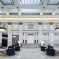 Luxurious hotel lobby with marble floor, white columns, central reception desk, skylight ceiling, crystal chandeliers, and purple seating.