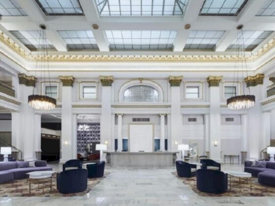 Luxurious hotel lobby with marble floor, white columns, central reception desk, skylight ceiling, crystal chandeliers, and purple seating.