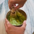 Hands whisking bright green matcha in a brown ceramic bowl with a bamboo whisk on a marble table, close-up view]