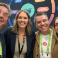 Four people smiling for a selfie at a conference, wearing lanyards, with a dark blue backdrop of colorful circular graphics behind them.