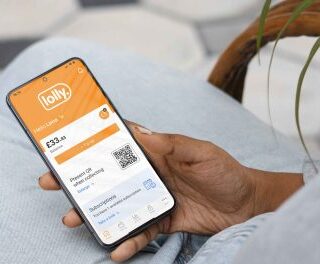 Close-up of a person in light jeans holding a smartphone displaying the Lolly app with a balance and a QR code on screen.
