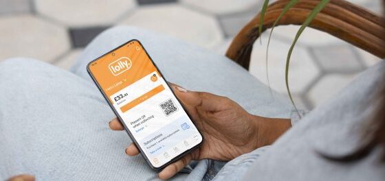 Close-up of a person in light jeans holding a smartphone displaying the Lolly app with a balance and a QR code on screen.