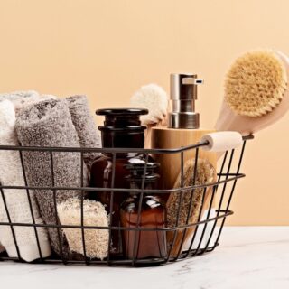 Black metal basket containing rolled towels, amber bottles, and wooden body brushes set against a peach background.