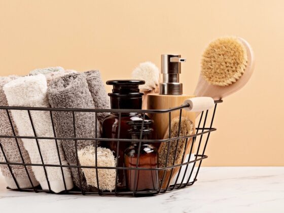 Black metal basket containing rolled towels, amber bottles, and wooden body brushes set against a peach background.