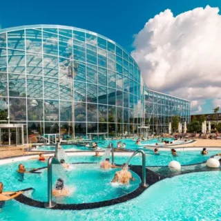 Outdoor pool next to a curved glass-domed building on a sunny day, with people swimming and lounging by the water.