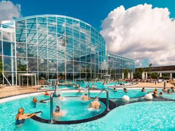 Outdoor pool next to a curved glass-domed building on a sunny day, with people swimming and lounging by the water.
