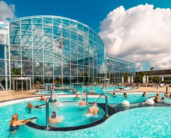 Outdoor pool next to a curved glass-domed building on a sunny day, with people swimming and lounging by the water.