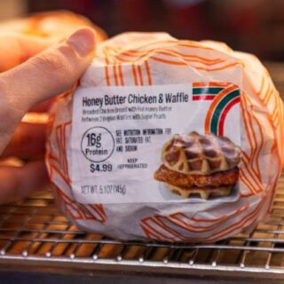Hand reaching to take a Honey Butter Chicken & Waffle sandwich from a store freezer shelf; white wrapper with orange stripes, label shows 16g protein and $4.99.