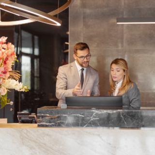Two professionals in business attire consult at a marble reception desk, with a tall pink floral arrangement on the left.