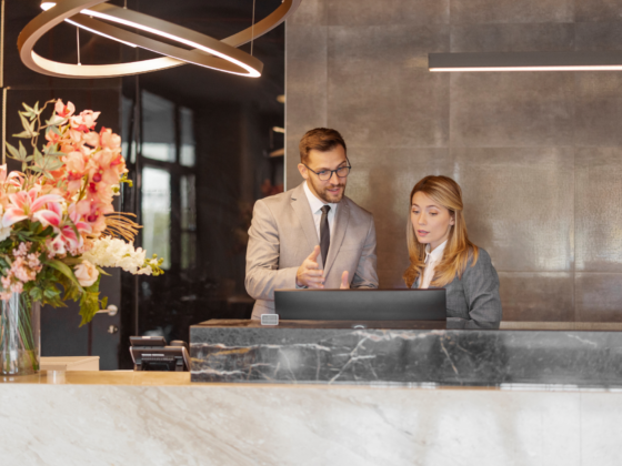 Two professionals in business attire consult at a marble reception desk, with a tall pink floral arrangement on the left.