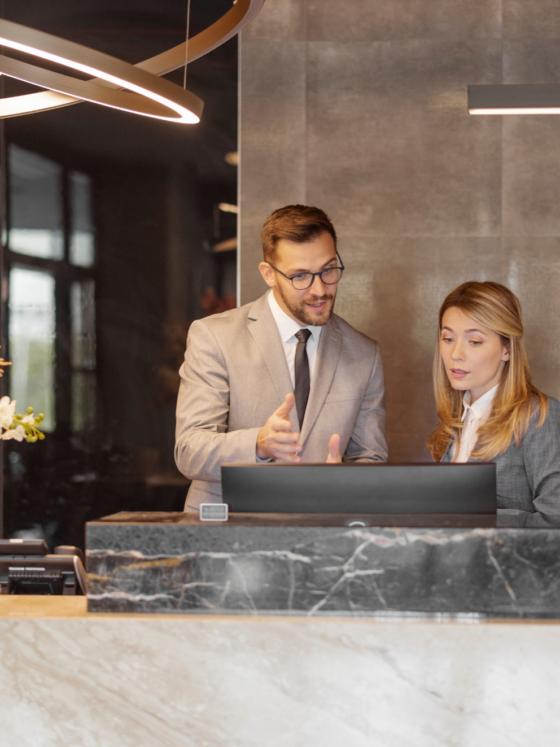 Two professionals in business attire consult at a marble reception desk, with a tall pink floral arrangement on the left.
