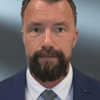 Close-up portrait of a man in a navy suit with a light gray patterned tie, looking at the camera, with a full beard.