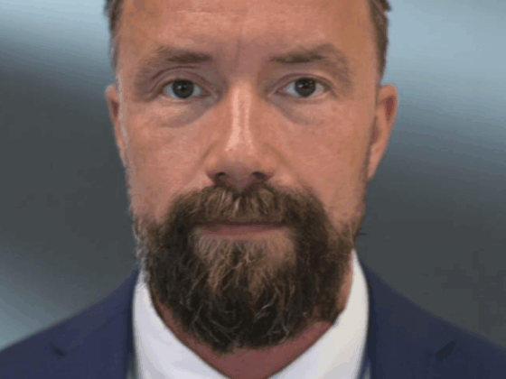 Close-up portrait of a man in a navy suit with a light gray patterned tie, looking at the camera, with a full beard.