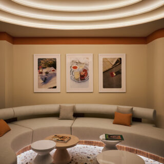 Curved beige sectional sofa with orange and gray cushions in a lounge, three drink-themed framed photos on the wall behind.