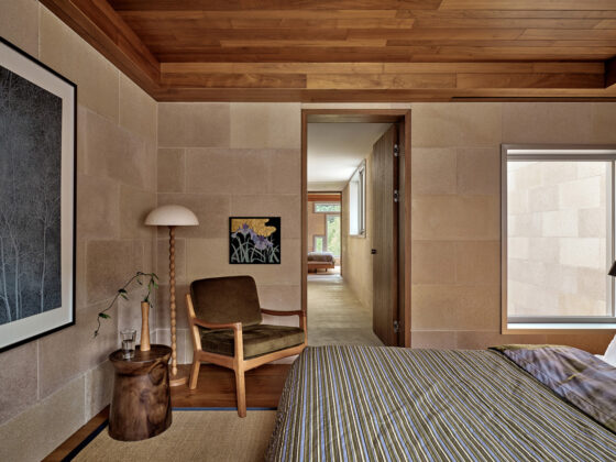 Bedroom corner with a mid-century chair, a wooden stump side table, and a tall floor lamp against stone walls and a striped duvet bed in the foreground.