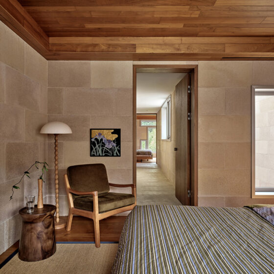 Bedroom corner with a mid-century chair, a wooden stump side table, and a tall floor lamp against stone walls and a striped duvet bed in the foreground.