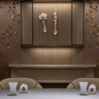 Elevator interior with two white spoon-like sculptures hanging on the door, flanked by floral wallpaper. Cresting wood trim above. Foreground shows a table with white vases.