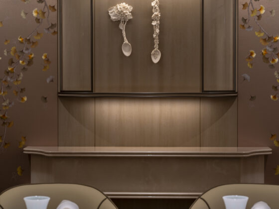 Elevator interior with two white spoon-like sculptures hanging on the door, flanked by floral wallpaper. Cresting wood trim above. Foreground shows a table with white vases.