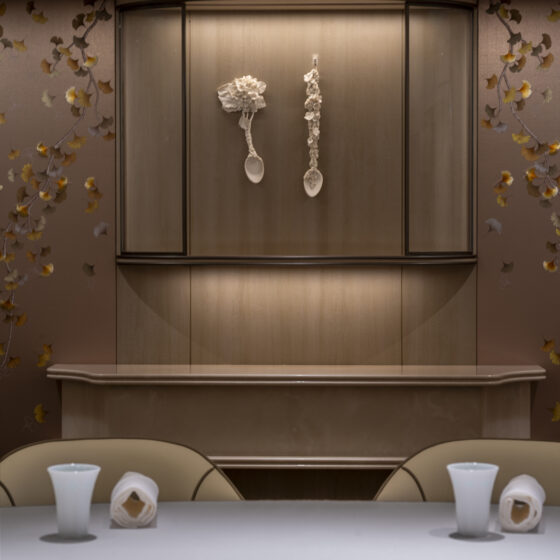 Elevator interior with two white spoon-like sculptures hanging on the door, flanked by floral wallpaper. Cresting wood trim above. Foreground shows a table with white vases.