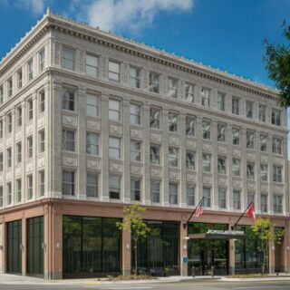 Five-story brick and stone office building with large ground-floor storefront windows, decorative cornice, and flags at the entrance on a city street.