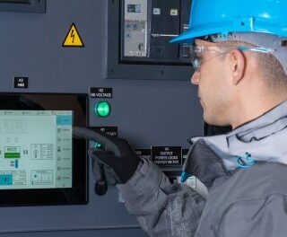 Worker in a blue hard hat and safety jacket uses a touchscreen control panel inside an electrical cabinet on site.