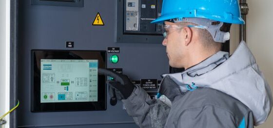 Worker in a blue hard hat and safety jacket uses a touchscreen control panel inside an electrical cabinet on site.