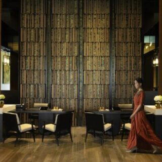 Woman in a red dress walks through a modern restaurant with dark wood paneling and a tall textured wall.