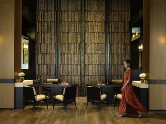Woman in a red dress walks through a modern restaurant with dark wood paneling and a tall textured wall.