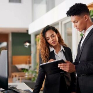 Two colleagues in business attire review a tablet together at a modern reception desk in a bright office area; they look focused and collaborative.
