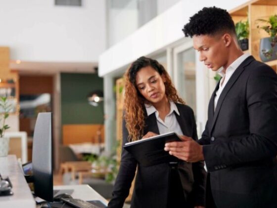 Two colleagues in business attire review a tablet together at a modern reception desk in a bright office area; they look focused and collaborative.