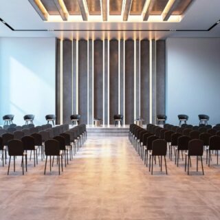 Modern conference room with many rows of black chairs facing a raised stage with a panel desk and vertical light panels, ready for a seminar. - or - Empty auditorium-style hall with rows of chairs toward a front stage and natural light from windows. - or - Bright, spacious lecture hall with floor-to-ceiling windows and a minimalistic stage at the front. - or - An event venue prepared for a presentation: rows of seating converging on a front table. - or - Large presentation room with numerous chairs, a front table, and decorative back wall lighting.